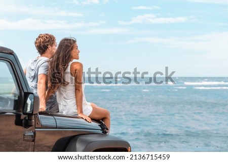 Car road trip travel couple tourists enjoying ocean view relaxing on hood of sports utility car. Happy Asian woman, man friends smiling on beach.