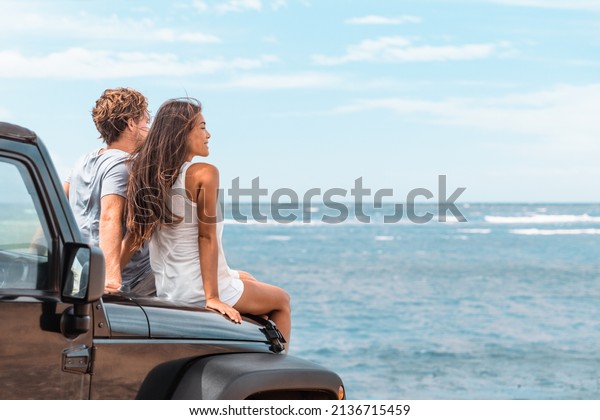 Car road trip travel couple tourists enjoying\
ocean view relaxing on hood of sports utility car. Happy Asian\
woman, man friends smiling on\
beach.