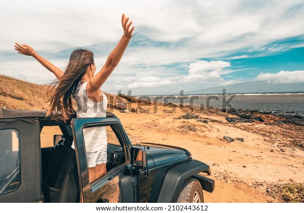 Car road trip travel fun\
happy woman tourist with open arms at ocean view from sports\
utility car driving on beach. Summer vacation adventure girl from\
the back.
