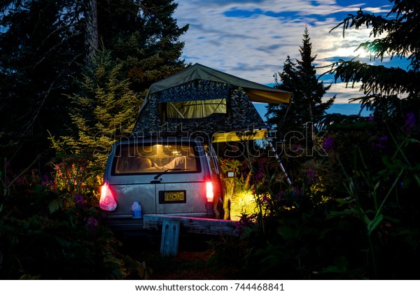 Car with rooftop tent in darkness with lights\
turned on in Alask