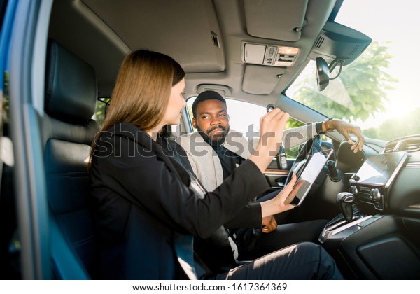 Car sale and rent, people concept. Happy African\
man and Caucasian woman car dealer with tablet computer sitting in\
new car. Woman salesmanager holds car keys and showing the rent\
contract on tablet