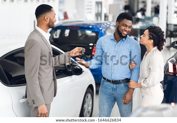 Car Sales. African\
American Family Buying Auto Taking Key From Dealer Standing In\
Luxury Dealership Store