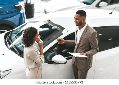 Car Sales Manager Showing Auto To Afro Lady Buyer Standing In Luxury Automobile Dealership Store. Buying Vehicle Concept