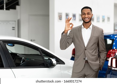 Car Sales. Smiling Black Salesman Gesturing Okay Standing In Luxury Auto Store. Free Space For Text