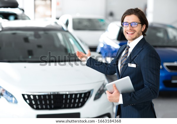 Car\
Sales. Smiling Salesman In Glasses Showing New Auto Standing In\
Automobile Dealership Showroom. Selective\
Focus