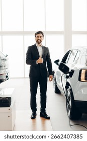 Car salesman is gesturing thumbs up while standing next to an electric car charging in a modern car dealership, expressing satisfaction and recommending the vehicle
