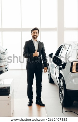 Car salesman is gesturing thumbs up while standing next to an electric car charging in a modern car dealership, expressing satisfaction and recommending the vehicle
