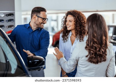 A car salesman is showing new cars to a couple. Car sales business. Manager talking to couple, showing them new auto at dealership shop. Young family selecting vehicle, looking at modern automobile  - Powered by Shutterstock