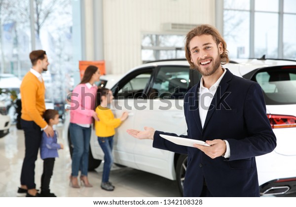 Car salesman with tablet and blurred family\
near auto on background