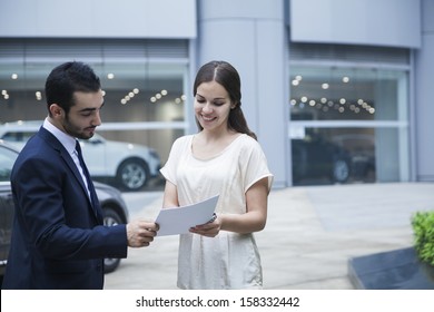 Car Salesman And Young Woman Looking Over Paperwork 