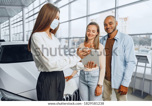 Car saleswoman wearing medical mask shows\
buyers couple something on digital\
tablet