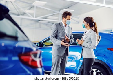 Car Seller Standing In Car Salon With A Customer And Showing On Tablet Car Performances And Specifications. They Both Have Face Masks On Faces Because It's Corona Outbreak.