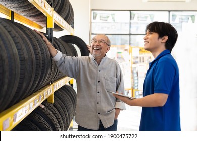 Car Service. Asian Salesman Recommend New Tire Wheel On Shelves Shelf To Senior Elderly Customer At The Auto Car Repair Shop. Specialist Mechanic And Customer Examining New Tire Wheel