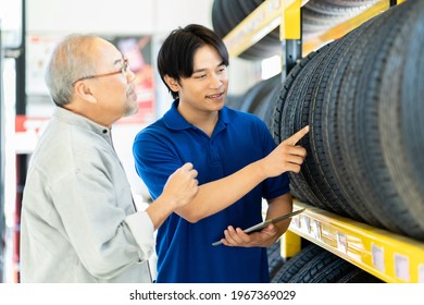 Car Service. Asian Salesman Recommend New Tire Wheel On Shelves Shelf To Senior Elderly Customer At The Auto Car Repair Shop. Salesman And Customer With New Tire Wheel At Car Workshop