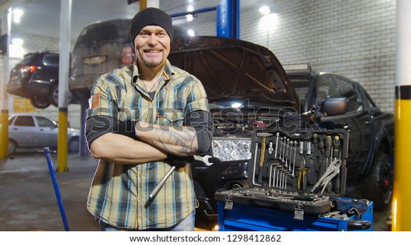 Car service. Brutal mechanic man standing by the\
car and smiling