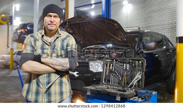 Car service. Brutal mechanic man standing by the\
car holding a wrench