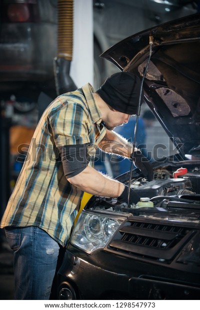 Car service. Brutal mechanic man\
standing by the car with open hood and working with a\
tool