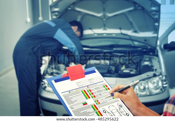 Car service. Holding the check list while car\
mechanic fixing the car in the background. Maintenance. Car broken\
down. Car break down. Car\
broke.
