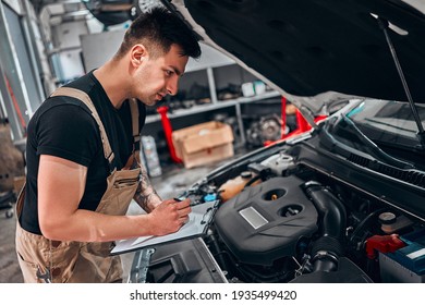Car service inventory. Mechanic in auto repair shop makes a note on a clipboard while checking on a car engine. Side view.