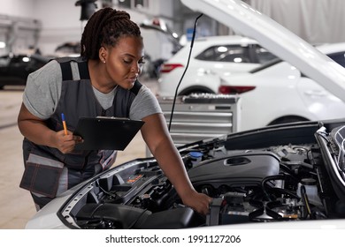 Car Service, Job And Profession Concept. Young African Female Worker In Gray Uniform With Clipboard And Pen Writing Over Auto Repair Shop On Background