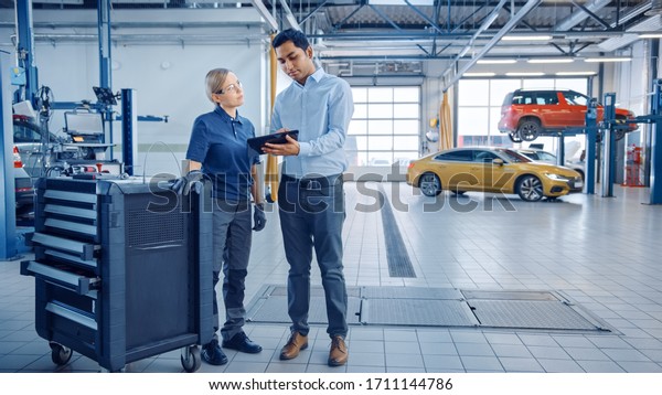 Car Service\
Manager and a Confident Female Mechanic Talk About Work Related\
Topics. They Use a Tablet Computer. Modern Clean Workshop with a\
Car on a Lift in the\
Background.