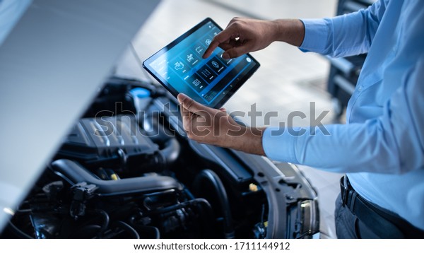 Car Service Manager or Mechanic Uses a Tablet\
Computer with a Futuristic Interactive Diagnostics Software.\
Specialist Inspecting the Vehicle in Order to Find Broken\
Components In the Engine\
Bay.