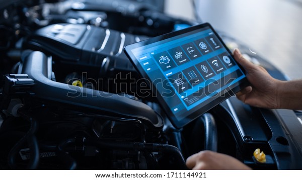 Car Service Manager or Mechanic Uses a Tablet\
Computer with a Futuristic Interactive Diagnostics Software.\
Specialist Inspecting the Vehicle in Order to Find Broken\
Components In the Engine\
Bay.