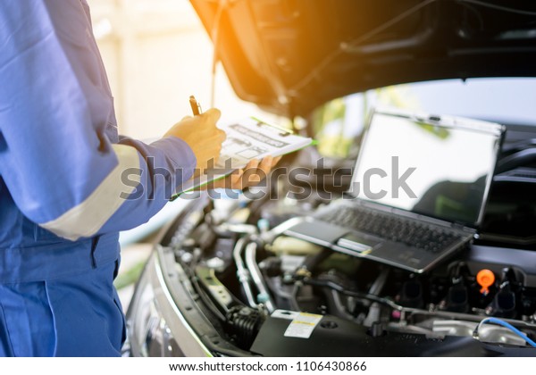 car service, repair, maintenance  concept - auto\
mechanic man or Smith writing to the clipboard at warehouse\
workshop, technician doing the checklist for repairs engine a car\
in the garage with laptop