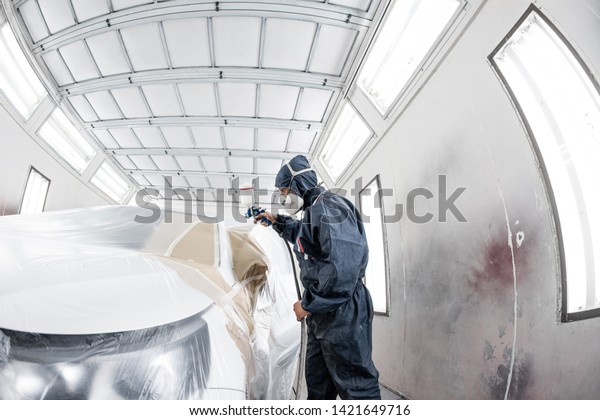 Car service station.\
Worker painting a white car in special garage, wearing costume and\
protective gear