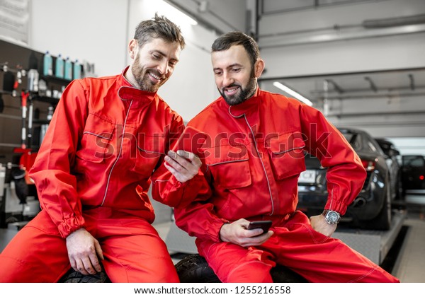 Car\
service workers in red uniform having a break sitting together with\
phone on the wheels at the tire mounting\
service