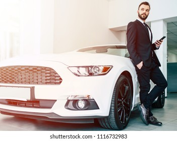 The car shows the character of a man. Confident success elegant business man in full suit and with phone in hand standing near new white sport cabriolet and looks away
