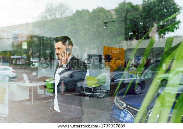 The car shows the character of a man.\
Confident success elegant business man in full suit and with phone\
in hand standing near new blue car looks\
away