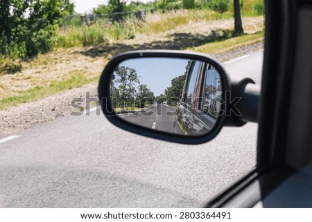 Car side mirror reflects an empty rural road on a sunny day. Travel safety and driving perspective background for road trip transport journey and quiet countryside transportation themes.