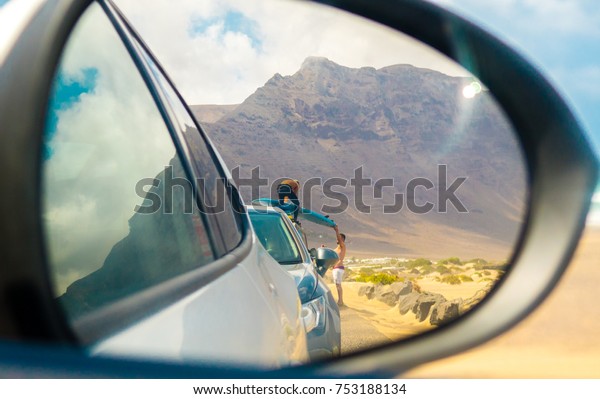 Car side view mirror photo of two men loading\
their surf boards onto their car at Famara Beach in Lanzarote,\
Canary Islands, Spain.