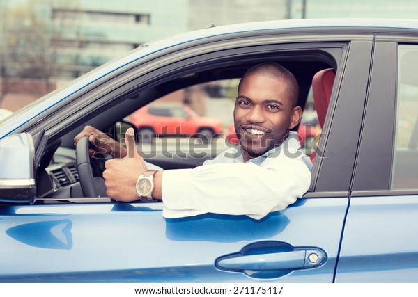 Car side window. Man driver happy smiling\
showing thumbs up driving sport blue car isolated outside parking\
lot background. Handsome young man excited about his new vehicle.\
Positive face expression