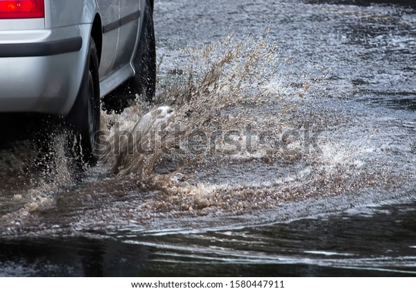 Car splashes\
through large puddle on flooded street. Motion car, rain, big\
puddle of water spray from\
wheels