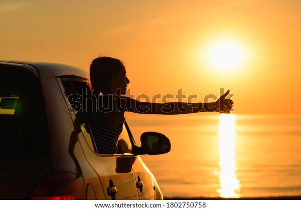 The car stands on the seashore at\
sunset. The girl climbed out of the car window and showed her thumb\
up. The joy of a girl from a trip to the sea by\
car.