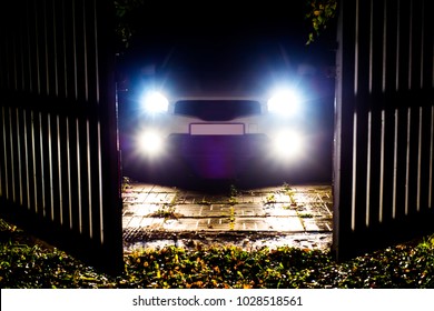 The Car Stands On The Territory Of The House At Night. Light Headlights Through The Fence