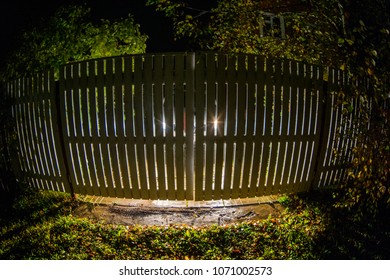 The Car Stands On The Territory Of The House At Night. Light Headlights Through The Fence