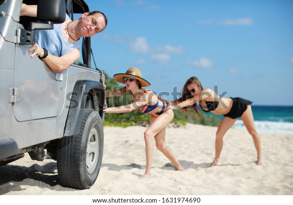 The car stuck\
in the sand.Two beauty and sexy women in bikini  pushing a off road\
car near sea on beach while man is emboldening\
them.Transportation,teamwork,funny\
concept.