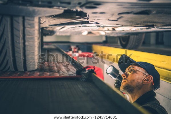 Car Suspension Issue.\
Caucasian Vehicle Mechanic with Flashlight Inspecting Car\
Undercarriage.