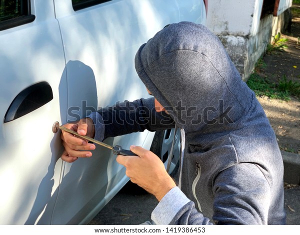 Car theft: man wearing a hood tries to break\
into a white car using skrewdriver as a key at daylight street in\
city. Car thief concept.