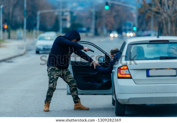 The car thief is pulling the car owner out of his\
car and trying to get the car while pointing a loaded gun at the\
drivers head.