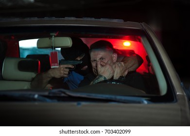 Car Thief Threatening Driver With The Gun. Male Robber Wearing Black Mask Sitting At The Back Seat And Pointing Gun At The Head Of Scared Owner. Police Light On The Background.