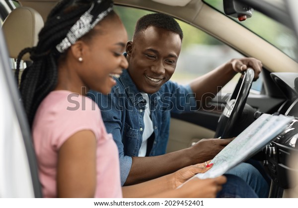 Car trip, travelling by auto concept. Happy\
millennial black woman showing her boyfriend destination to travel\
on paper map, sitting at front seats in automobile, discussing\
future journey, side view