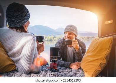 Car trunk view of chatting couple dressed warm knitted clothes enjoying gas stove prepared coffee and mountain lake view. Cozy early autumn couple auto traveling concept image. - Powered by Shutterstock