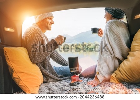 Car trunk view of chatting couple dressed warm knitted clothes enjoying gas stove prepared coffee and mountain lake view. Cozy early autumn couple auto traveling concept image.  