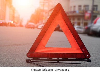 Car Warning Triangle On The Road Against The City In The Evening. Red Warning Triangle With A Car.