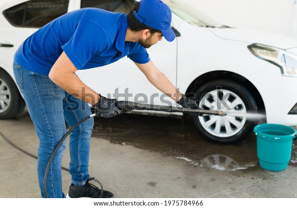 Car wash and auto detail service. Hispanic male\
worker with gloves using a water hose to finish washing a clean\
white vehicle of a customer