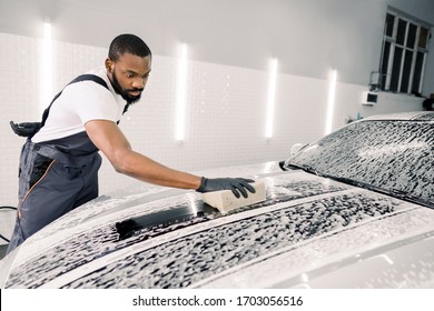 Car Wash And Clean With Shampoo And Sponge. Young Concentrated African American Man Worker, Wearing Gray Overalls And Black Gloves Washing Soapy Car Hood With Sponge On A Car Wash.